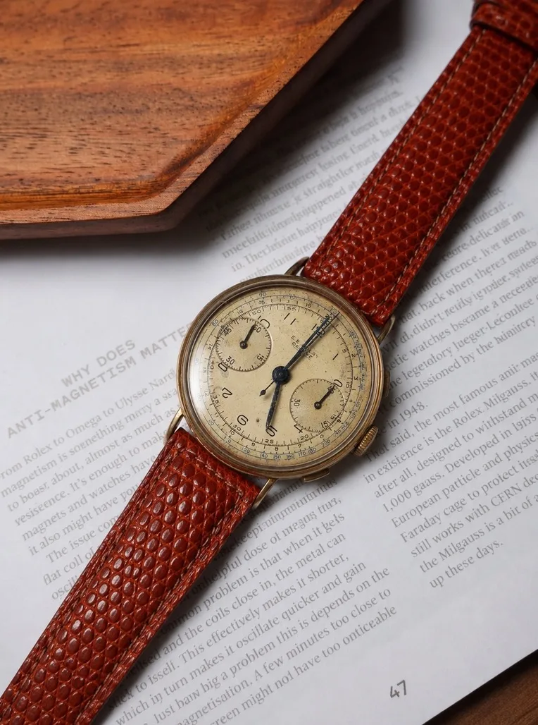 A vintage gold chronograph on a brown lizard strap, resting on an open watch book